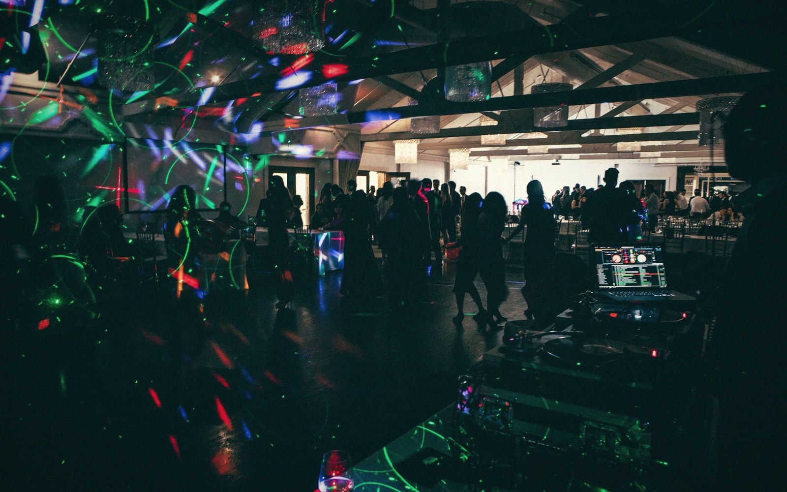 DJ booth and dance floor lit by colorful lasers as a crowd gathers under a wooden-beamed ceiling